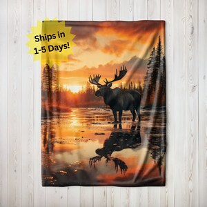 May include: A blanket depicting a moose in a lake at sunset, with warm orange and red tones. The moose's reflection is in the water. Trees are in the background. The text "Ships in 1-5 Days!" is in the upper left corner. The blanket is displayed against a wooden backdrop.