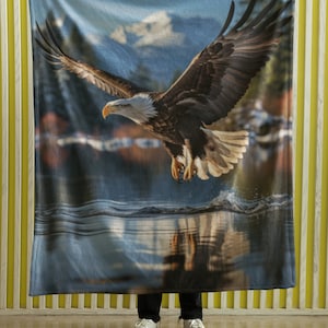 May include: A blue fleece blanket featuring a printed image of a bald eagle in flight over a lake. The eagle is brown and white with a yellow beak. The lake is blue and the sky is blue with white clouds.