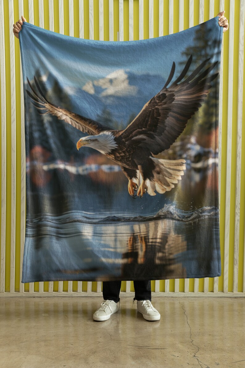 May include: A blue fleece blanket featuring a printed image of a bald eagle in flight over a lake. The eagle is brown and white with a yellow beak. The lake is blue and the sky is blue with white clouds.