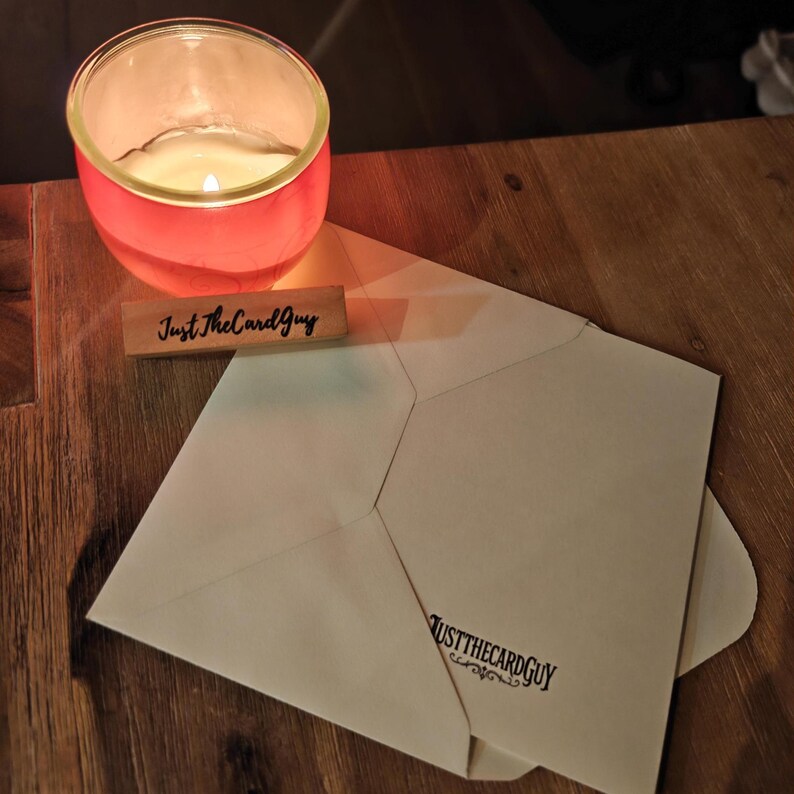 May include: A lit candle in a red glass votive, a wooden sign with the text "Just The Card Guy", and a cream-colored envelope with the text "JUSTTHECARDGUY" printed on it. The items are on a wooden surface.