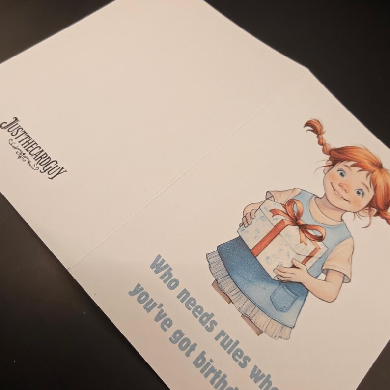 May include: Greeting card with an illustration of a girl holding a gift. The girl has red pigtails and is wearing a blue dress. Text on the card reads "Who needs rules when you've got birthdays?" The card is from "JUSTTHECARDGUY".