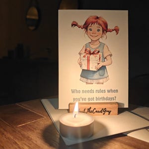 May include: A birthday card featuring a cartoon girl with red pigtails holding a gift. The card reads, "Who needs rules when you've got birthdays?" A lit candle sits in front of the card, with a wooden holder that says "Just TheCardGuy."