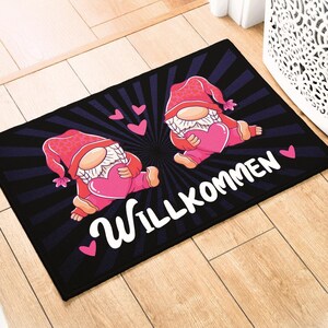 Può includere: Un tappetino da porta nero con un messaggio di benvenuto bianco che dice "Willkommen". Il tappetino presenta due gnomi rosa che tengono cuori rosa.