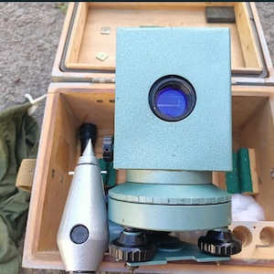 May include: A light green surveying instrument with a circular blue lens, a metallic plumb bob with a pointed tip, and black adjustment knobs, all housed within a rectangular wooden storage box with a green fabric pouch nearby.