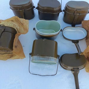 Puede incluir: Una colección de kits de cocina de metal en color verde oliva, algunos con tapas cerradas y otros abiertos que revelan un interior claro. Varios kits cuentan con asas de metal y mecanismos de cierre, con algunos artículos envueltos en papel marrón.