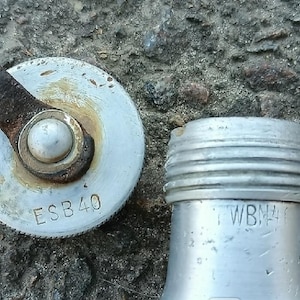 Puede incluir: Una tapa metálica circular de color plateado con un pestillo marrón oxidado y el texto ESB40, junto a un cuello de botella metálico plateado con crestas roscadas y el texto parcialmente visible TWBN40, ambos con acabado metálico.