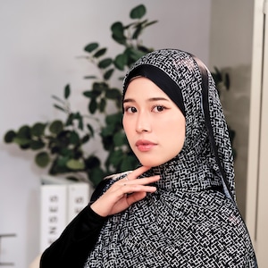 May include: A black and white patterned hijab. The hijab features a geometric design and is worn with a black top. The image is a medium shot, with the subject looking towards the camera. The background is blurred, with hints of greenery and neutral tones.