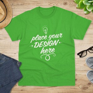 Könnte beinhalten: Ein grünes T-Shirt mit dem Text "place your design here" in Weiß. Das Shirt liegt flach auf einer Holzoberfläche mit einem Strohhut, Jeans und Turnschuhen.