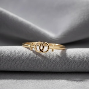 Peut inclure: Une bague dorée ornée d'un double symbole féminin. La bague est présentée sur un tissu gris texturé, mettant en valeur ses détails délicats et son éclat. Le design de la bague est un symbole de la fierté lesbienne.