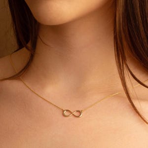 May include: A delicate gold-colored necklace featuring an infinity symbol pendant. The pendant is suspended from a thin chain, creating a minimalist and elegant design. The necklace is worn around the neck.