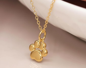 Könnte beinhalten: Eine goldfarbene Halskette mit einem Pfotenabdruck-Anhänger. Der Anhänger hat ein detailliertes Pfotenabdruck-Design mit fünf abgerundeten Pads. Die Kette ist zart und goldfarben und schafft ein schlichtes, aber elegantes Schmuckstück.