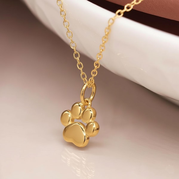 Könnte beinhalten: Eine goldfarbene Halskette mit einem Pfotenabdruck-Anhänger. Der Anhänger hat ein detailliertes Pfotenabdruck-Design mit fünf abgerundeten Pads. Die Kette ist zart und goldfarben und schafft ein schlichtes, aber elegantes Schmuckstück.