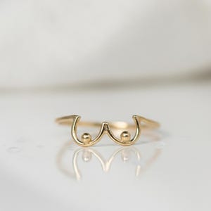 Può includere: Un anello dorato dal design unico. L'anello presenta una rappresentazione stilizzata di un paio di seni, con due forme arrotondate e linee curve. L'anello è posto su uno sfondo bianco.