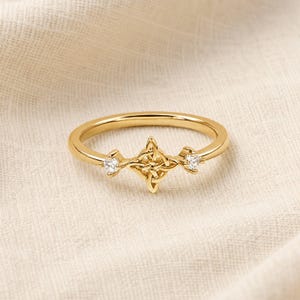 May include: A gold ring featuring a central knot design flanked by two small, clear, round-cut stones. The band is smooth and polished, with the knot motif positioned horizontally across the top, creating a symmetrical and detailed appearance.