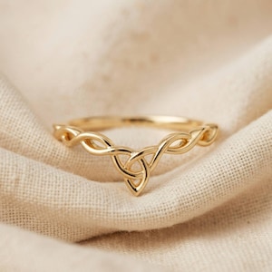 May include: A gold ring featuring a V-shaped band with a central triquetra knot design. The metal has a polished finish, and the band consists of intertwined strands that form a continuous, symmetrical pattern across the front of the ring.