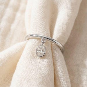 May include: A silver ring featuring a dangling oval gemstone. The ring is displayed against a soft, off-white fabric. The gemstone is clear and sparkling, enhancing the ring's simple, elegant design. A delicate piece of jewellery.