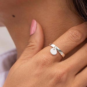 Könnte beinhalten: Ein silberner Ring mit einem kleinen, runden Anhänger. Der Ring wird am Finger getragen, wobei der Anhänger frei hängt. Der Ring hat eine polierte Oberfläche, die das Licht reflektiert. Der Schmuck ist schlicht und elegant.