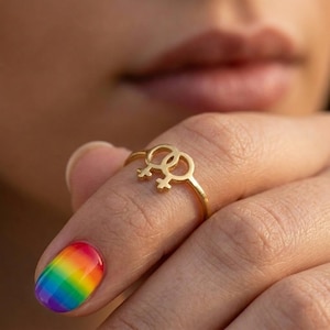 Op de afbeelding: Een goudkleurige ring met een dubbel vrouwelijk symboolontwerp. De ring wordt gedragen aan een vinger met een regenboogkleurige nagel. De ring is een symbool van lesbische trots en is een sieraad.