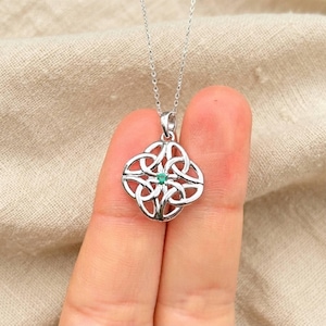 Pode incluir: Um colar de tom prateado com um pendente de design de nó simétrico e intrincado. O centro do pendente está engastado com uma pequena pedra preciosa redonda de cor verde vibrante. O pendente pende de uma fina corrente de metal prateado.