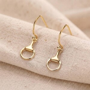 Könnte beinhalten: Ein Paar goldfarbene Ohrhänger mit einem Trensen-Design. Jeder Ohrring besteht aus einem geschwungenen Haken, der mit einem kleinen Metallsteg und einem kreisförmigen Ring verbunden ist, was für ein poliertes, metallisches Finish sorgt.
