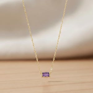 May include: A gold necklace featuring a rectangular, purple gemstone pendant. The pendant is set in a gold bezel and suspended from a delicate gold chain. The necklace is presented on a light-coloured wooden surface.