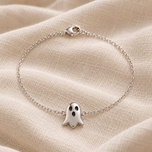 Op de afbeelding: Een zilverkleurige metalen kettingarmband met een centrale bedel in de vorm van een gestileerde geest met twee ovale ogen en een open mond. De armband is voorzien van een kleine karabijnsluiting aan de bovenkant die de fijne schakelketting verbindt.