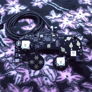 May include: A black PhobGCC 2.0.5 printed circuit board for a game controller featuring two white analog sticks, gold contact pads, a ghost icon, and a black wired cable with a connector plug attached to the top of the board.