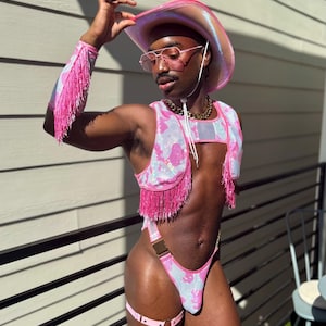 May include: A pink and white sequined cowboy hat, matching vest, and thong bodysuit with pink fringe. The outfit includes pink arm cuffs and garter straps. The model is wearing pink-tinted glasses and a gold chain.