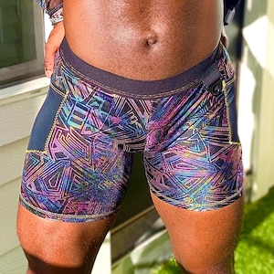 May include: Close-up of colourful, geometric-patterned shorts. The shorts feature a dark waistband and side panels with gold stitching. The design includes a vibrant mix of purple, blue, and yellow lines, creating a holographic effect. The shorts are made of a shiny material.
