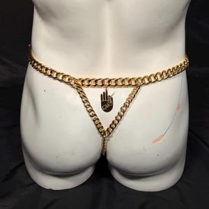 Könnte beinhalten: Ein goldfarbener Metallketten-String mit einer horizontalen Taillenkette, die mit einer vertikalen Mittelkette verbunden ist. Ein kleiner, runder Anhänger hängt in der Mitte und zeigt eine Grafik einer Hand mit gespreizten Fingern und den Buchstaben OVR.