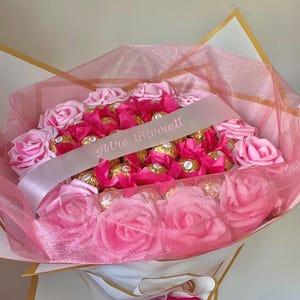 Puede incluir: Un ramo de rosas rosas y bombones envueltos, acentuado con tul rosa y una cinta blanca que dice "Mrs Barrett". El ramo est&aacute; envuelto en papel blanco y dorado.