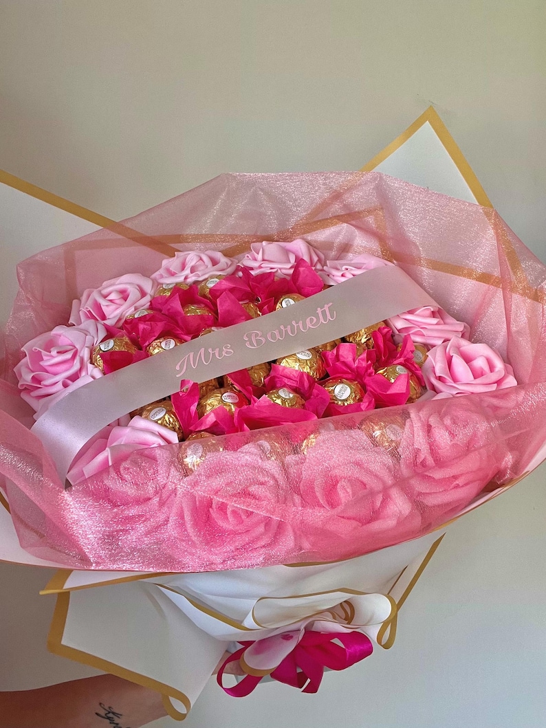 Puede incluir: Un ramo de rosas rosas y bombones envueltos, acentuado con tul rosa y una cinta blanca que dice "Mrs Barrett". El ramo est&aacute; envuelto en papel blanco y dorado.