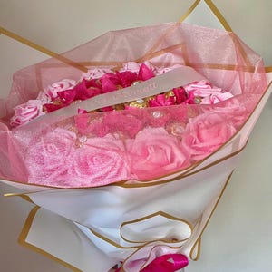 Puede incluir: Un ramo de rosas rosas y bombones, envuelto en papel blanco y ribeteado en dorado. El ramo est&aacute; envuelto en una red rosa y tiene una cinta con el texto "Mrs. Garrett". Una cinta rosa brillante est&aacute; atada en la base.