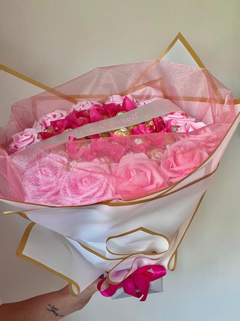 Puede incluir: Un ramo de rosas rosas y bombones, envuelto en papel blanco y ribeteado en dorado. El ramo est&aacute; envuelto en una red rosa y tiene una cinta con el texto "Mrs. Garrett". Una cinta rosa brillante est&aacute; atada en la base.