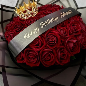 May include: A bouquet of deep red roses featuring a gold-toned crown with clear crystals resting on top. A black ribbon is draped across the flowers with the gold text Happy Birthday Ashanti! printed on it, all wrapped in dark translucent paper.