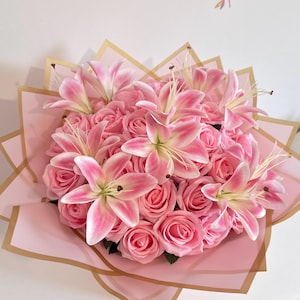 May include: A bouquet of pink roses and white lilies with pink-tipped petals, arranged in a circular cluster. The flowers are wrapped in light pink paper with gold-coloured edges, forming a star-shaped border around the floral arrangement.