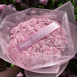 Könnte beinhalten: Ein kreisförmiger Strauß aus hellrosa Kunstrosen, eingewickelt in durchscheinenden rosa Organzastoff. Ein hellrosa Band ist diagonal über die Mitte der Rosen drapiert und trägt den weißen Aufdruck Happy Birthday Diane.