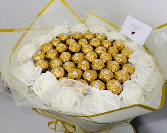 May include: A bouquet of white roses and gold-wrapped chocolates. The chocolates are arranged in the center, surrounded by the white roses. The bouquet is wrapped in white and gold paper, with a small card that reads "Blooming Gifts".