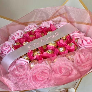 Puede incluir: Un arreglo floral rosa con rosas rosas y bombones envueltos. Una cinta rosa claro con el texto "Mrs Barrett" est&aacute; drapeada sobre el arreglo. El ramo est&aacute; envuelto en papel de seda rosa.