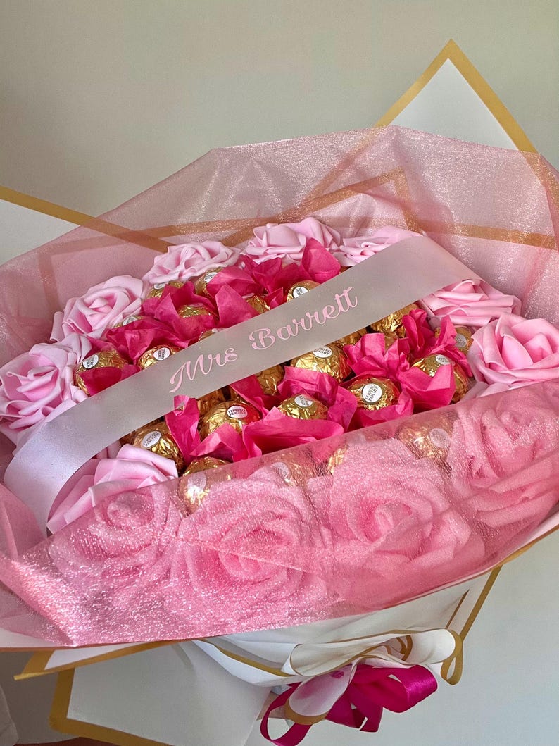 Puede incluir: Un arreglo floral rosa con rosas rosas y bombones envueltos. Una cinta rosa claro con el texto "Mrs Barrett" est&aacute; drapeada sobre el arreglo. El ramo est&aacute; envuelto en papel de seda rosa.