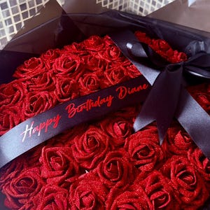 Könnte beinhalten: Ein herzförmiges Arrangement aus roten Rosen, präsentiert in einer schwarzen Box und mit schwarzem Tüll umwickelt. Ein schwarzes Band mit dem roten Schriftzug "Happy Birthday Diane" ist über die Rosen drapiert.