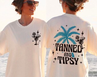 May include: White t-shirt with a black graphic of a cocktail glass with a lime wedge and lightning bolt. The back of the shirt has a blue palm tree with the text "Tanned and Tipsy" in black.