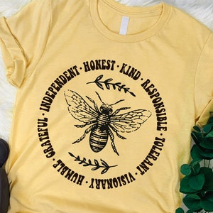 Peut inclure: T-shirt jaune clair avec un dessin noir d'abeille entouré des mots : Indépendant, Honnête, Gentil, Responsable, Tolérant, Visionnaire, Humble, Reconnaissant. Le t-shirt a des manches courtes et un col rond.