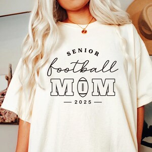 May include: Cream-colored t-shirt with the text "SENIOR football MOM 2025" in black. The word "football" is in cursive, and "MOM" is in bold, block letters with a football graphic. The shirt is a casual, oversized fit.