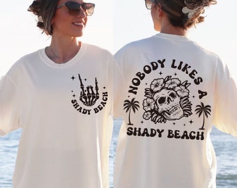 May include: White t-shirt with a black skull and flowers graphic on the back and the text "Nobody Likes a Shady Beach". The front of the shirt has a black skeleton hand making a rock and roll sign with the text "Shady Beach" in a circle.