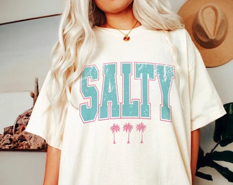 May include: A white t-shirt with the word "SALTY" printed in blue and pink with palm trees underneath.