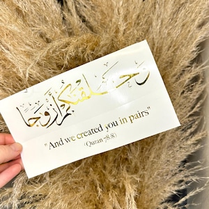 Könnte beinhalten: Goldfolie-Kalligraphie auf weißem Hintergrund mit dem arabischen Text "رحمن زوجا" und dem englischen Text "And we created you in pairs" (Koran 78:8).