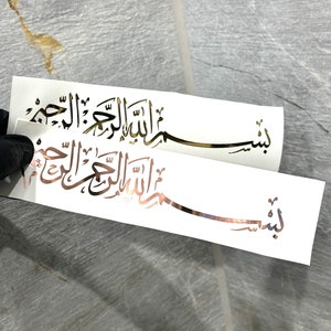 Könnte beinhalten: Zwei weiße Aufkleber mit goldener arabischer Kalligraphie. Der obere Aufkleber lautet "Bismillah" und der untere Aufkleber lautet "Allah" in einer dekorativen Schrift.