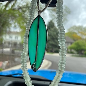 May include: A stained glass surfboard pendant in shades of green and teal, framed with dark metal. It hangs from a string of clear, rounded beads, suspended by a natural fiber cord. The pendant is a decorative car accessory.