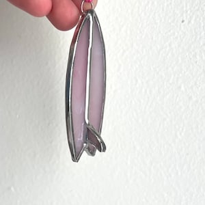 May include: A pink stained glass surfboard ornament with a silver frame. The ornament has a loop at the top for hanging.